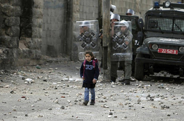 A young girl, unconcerned by the violence around her, makes her way to school past Israeli troops with protective shields.